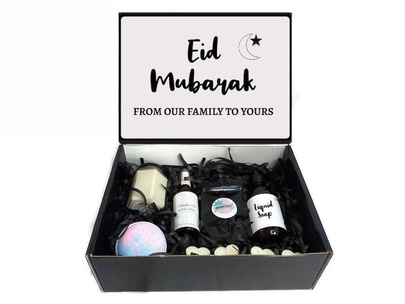 May include: A black gift box with a white lid. The lid has the text "Eid Mubarak" and a crescent moon and star design. The box is filled with black shredded paper and contains a candle, bath bomb, bottle of liquid soap, and a small bottle of room spray. The box is a gift for Eid.