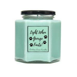 May include: A light blue hexagonal candle jar with a black lid and a white label that reads "Light When George Farts!" with paw prints. The label also includes the text "Lindsay Lucas Candles".