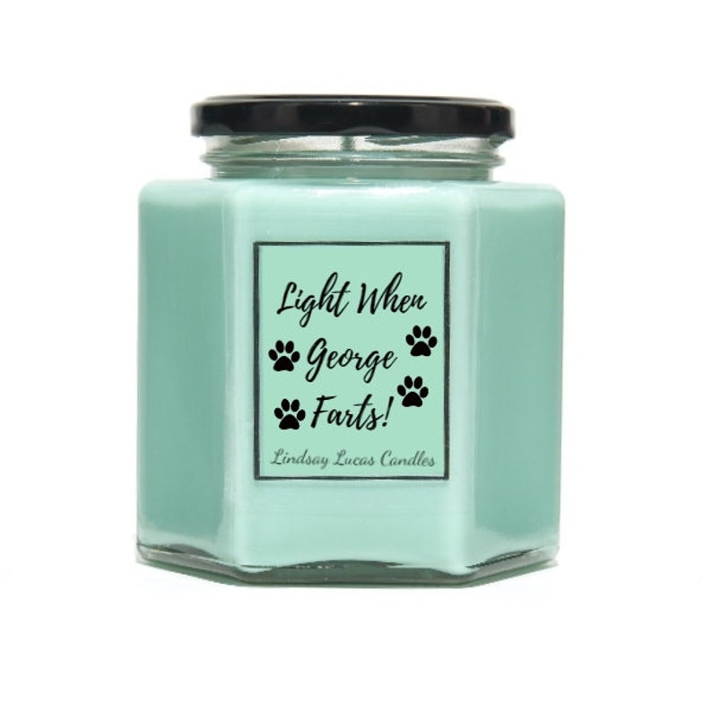 May include: A light blue hexagonal candle jar with a black lid and a white label that reads "Light When George Farts!" with paw prints. The label also includes the text "Lindsay Lucas Candles".