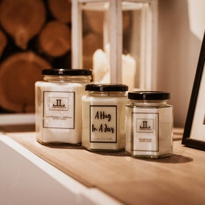 May include: Three glass jar candles with black lids and white labels. The labels read "Lavender & Vanilla", "A Hug In A Day", and "Lily & Rose". The candles are on a wooden shelf with a framed picture and a decorative lantern in the background.