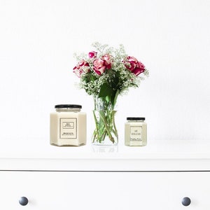 May include: Two candles in hexagonal glass jars with black lids, one large and one small, sit on a white surface. A vase of pink roses and white filler flowers is between them. The large candle has a label with the initials "JL".