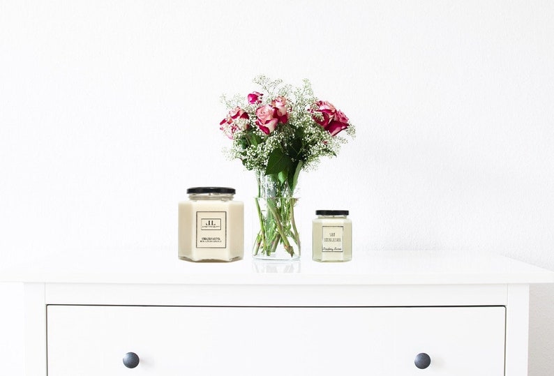 May include: Two candles in hexagonal glass jars with black lids, one large and one small, sit on a white surface. A vase of pink roses and white filler flowers is between them. The large candle has a label with the initials "JL".