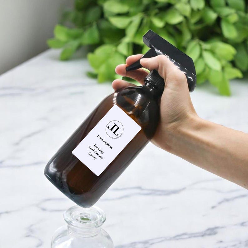 May include: A brown glass spray bottle with a black spray nozzle is held in a hand. A white label on the bottle reads "Lemongrass Ironing Anti Crease Spray" with a logo. The bottle is partially over a glass jar.