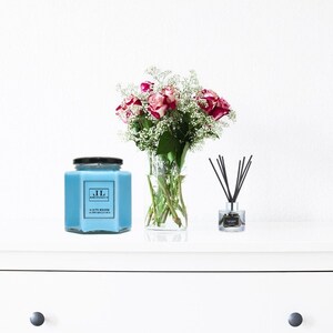 May include: A white dresser with a drawer and a blue glass candle jar with a black lid and a label that says "JL" and "CLAY & SAGE" on a white surface. There is a bouquet of pink roses and white baby's breath in a clear glass vase and a glass diffuser with black reeds on the surface.