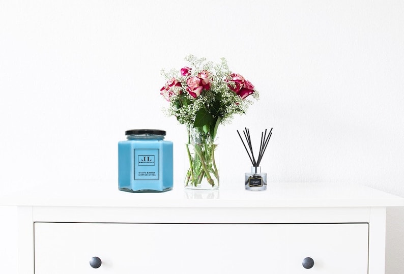 May include: A white dresser with a drawer and a blue glass candle jar with a black lid and a label that says "JL" and "CLAY & SAGE" on a white surface. There is a bouquet of pink roses and white baby's breath in a clear glass vase and a glass diffuser with black reeds on the surface.