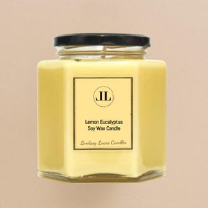 May include: A hexagonal glass jar candle with a black lid. The candle is filled with yellow wax and has a label that reads "Lemon Eucalyptus Soy Wax Candle" with the brand name "Lindsay Lucas Candles".