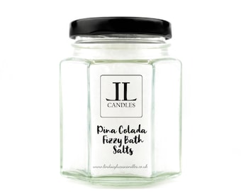 May include: A glass jar with a black lid filled with white bath salts. The jar has a label that says "Pina Colada Fizzy Bath Salts" and "JL Candles" with the website address "www.lindsayjacascandles.co.uk".