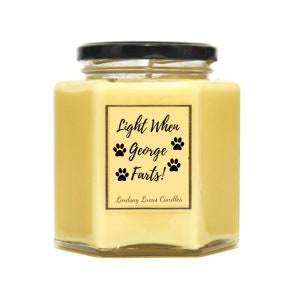 May include: A yellow hexagonal glass candle jar with a black lid. The label reads "Light When George Farts!" with paw prints and the text "Lindsay Lucas Candles".