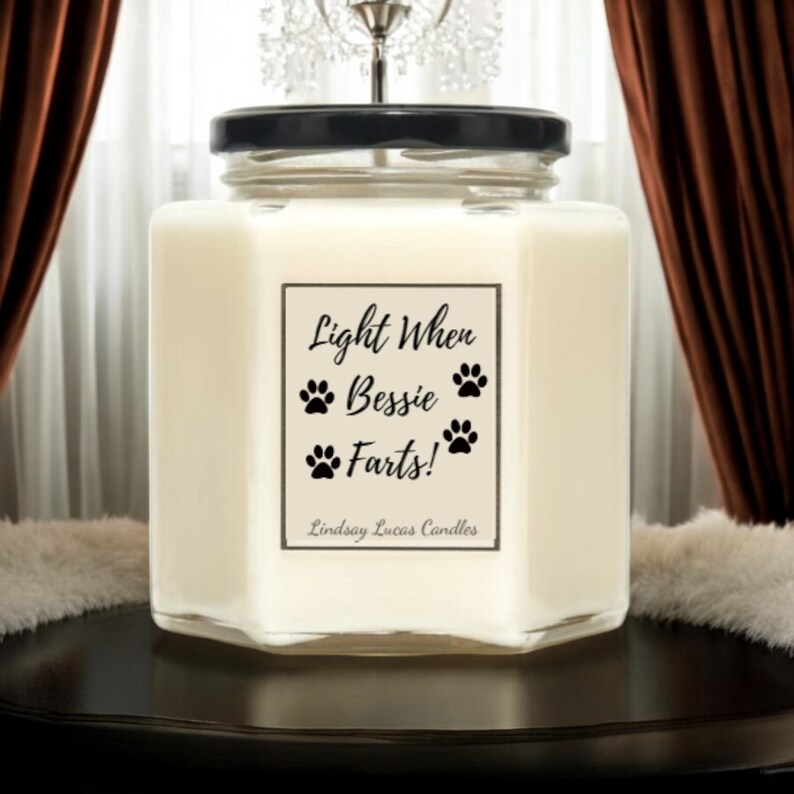 May include: A white candle in a clear glass jar with a black lid. The label reads "Light When Bessie Farts!" with paw prints around the text. The label also includes the text "Lindsay Lucas Candles".