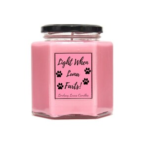May include: Pink glass jar candle with a black lid and a label that reads "Light When Luna Farts!" with paw prints. The label also includes the text "Lindsay Lucas Candles".