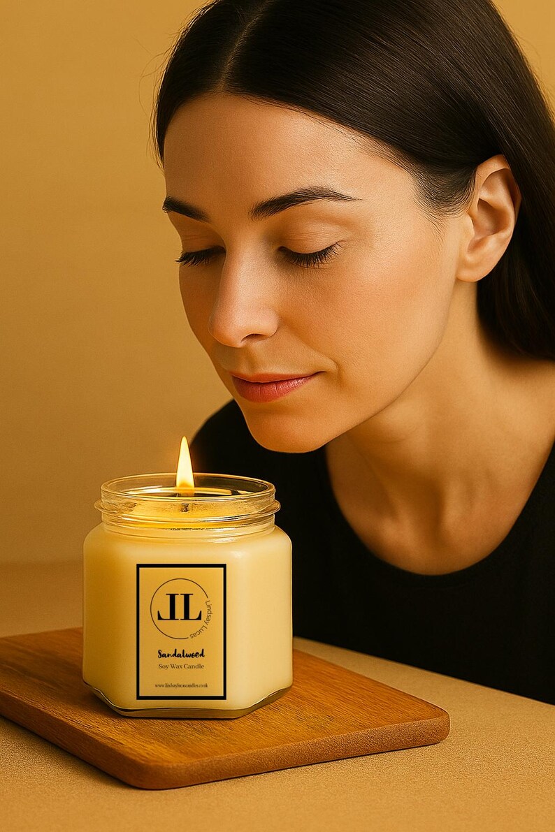 May include: A lit soy wax candle in a clear glass jar with a black and white label that reads "Sandalwood". The candle sits on a wooden surface. The flame is bright and the candle is yellow.