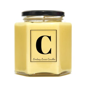 May include: A yellow candle in a hexagonal glass jar with a black lid. The candle has a label with the letter "C" in black and the text "Lindsay Lucas Candles".