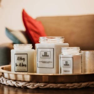 May include: A collection of scented candles in glass jars, displayed on a gold tray. The candles have white wax and labels with text. One reads "A Hug In A Jar", another "Lavender & Vanilla". The tray sits on a woven mat.