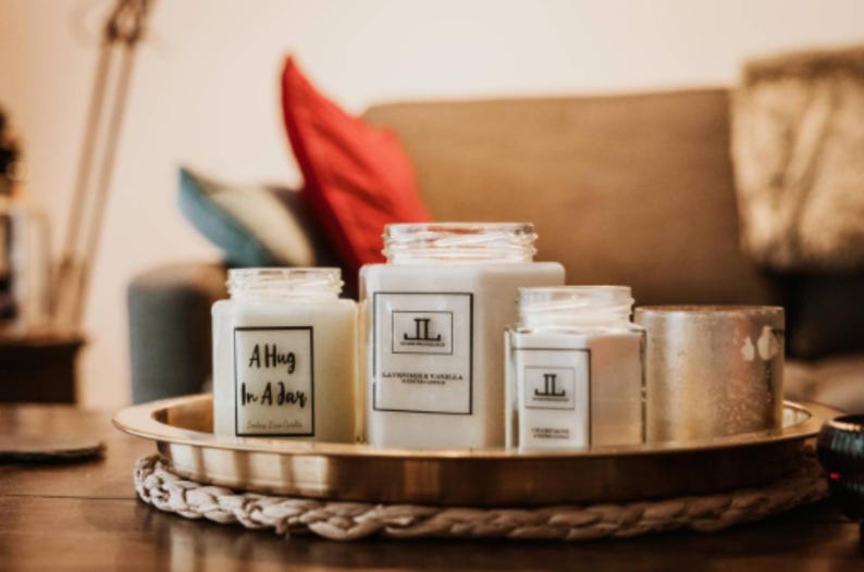 May include: A collection of scented candles in glass jars, displayed on a gold tray. The candles have white wax and labels with text. One reads "A Hug In A Jar", another "Lavender & Vanilla". The tray sits on a woven mat.