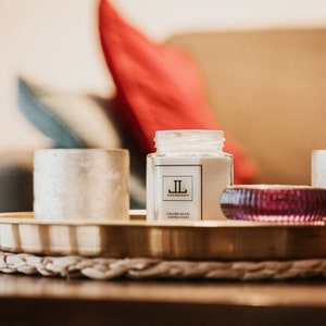 May include: A decorative tray with candles and a small glass jar candle. The jar candle has a label with the letters "JL" and the words "Champagne Scented Candle". A small, ribbed, purple glass candle holder is also on the tray.