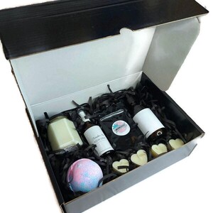 May include: A black gift box filled with bath and body products. The box contains a candle, two bottles of room spray, a bath bomb, and two heart-shaped bath melts. The box is filled with black shredded paper.