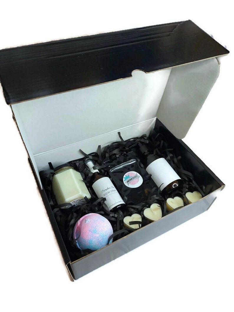 May include: A black gift box filled with bath and body products. The box contains a candle, two bottles of room spray, a bath bomb, and two heart-shaped bath melts. The box is filled with black shredded paper.