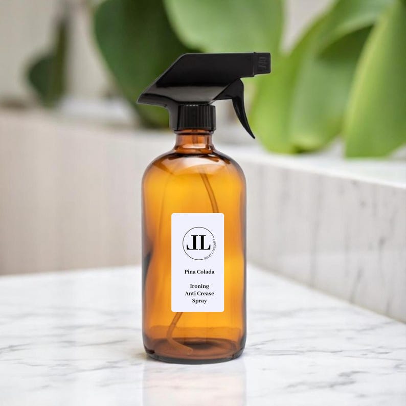 May include: An amber glass spray bottle with a black nozzle. The label reads "Pina Colada Ironing Anti Crease Spray" with the initials "JL" in a circle. The bottle is on a white marble surface.