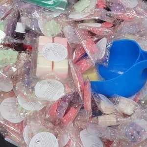 May include: A large black bin filled with small, individually packaged wax melts. The wax melts are in a variety of colors and scents. A blue plastic scoop is being used to remove the wax melts from the bin.