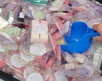 May include: A large black bin filled with small, individually packaged wax melts. The wax melts are in a variety of colors and scents. A blue plastic scoop is being used to remove the wax melts from the bin.