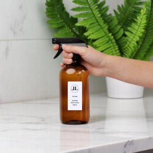 May include: A brown glass spray bottle with a black trigger, labelled with the text "Savage Ironing Anti Crease Spray". The bottle is held in front of a white background with a green plant.