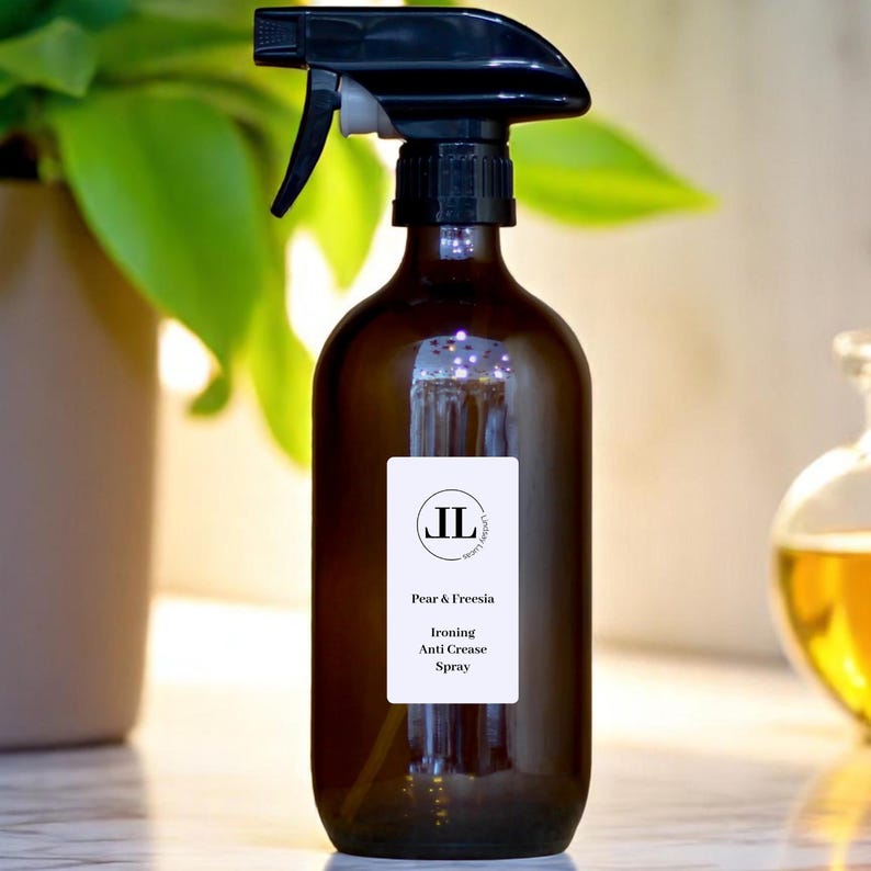 May include: A brown glass spray bottle with a black spray nozzle. The bottle has a white label with the text "Pear & Freesia Ironing Anti Crease Spray". The background includes green leaves and a blurred glass bottle.