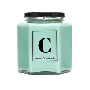 May include: A light green hexagonal glass candle jar with a black lid. The jar has a white label with the letter "C" in black and the text "Lindsay Lucas Candles".