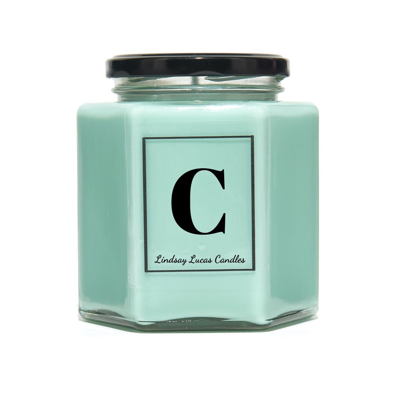 May include: A light green hexagonal glass candle jar with a black lid. The jar has a white label with the letter "C" in black and the text "Lindsay Lucas Candles".