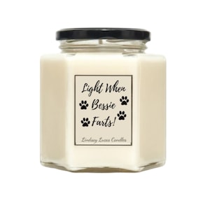 May include: A white candle in a glass jar with a black lid. The label reads "Light When Bessie Farts!" with paw prints. The candle is made by Lindsay Lucas Candles.
