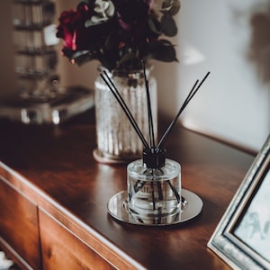Op de afbeelding: Een heldere glazen diffuserfles met zwarte rietjes en een zwart label met de tekst "Reed Diffuser". De fles staat op een zilveren dienblad op een houten oppervlak. Op de achtergrond staat een vaas met bloemen.