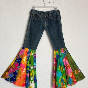 May include: Blue denim jeans with colorful floral patchwork bell bottoms. The patchwork includes vibrant hues of pink, orange, green, and yellow, creating a retro, bohemian style. The jeans are hanging on a clear hanger against a white wall.