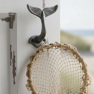 May include: A dark grey metal wall hook shaped like a whale tail, mounted on a white wooden surface. A circular wooden frame with a light tan woven fishing net is suspended from the hook, featuring a textured finish and visible screw holes.