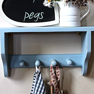 Può includere: Un ripiano in legno blu chiaro con quattro ganci e un cartello in ardesia che dice "Pegs". Il ripiano ha un bordo bianco ed è montato su una parete. Ci sono due strofinacci appesi ai ganci.