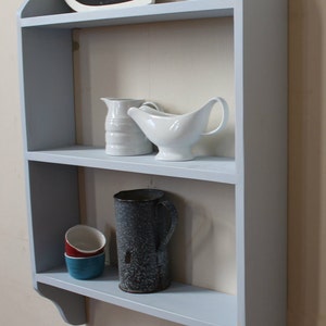 May include: A light blue wooden wall shelf with two shelves. The top shelf has a heart-shaped chalkboard sign that says "6 sizes available". The bottom shelf has a gray metal pitcher, two white ceramic pitchers, and three stacked ceramic cups in red, blue, and white.
