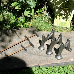 Può includere: Set di quattro cavalletti per camino in ghisa con un designo decorativo. I cavalletti sono di colore grigio scuro e sono disposti in fila su una superficie di pietra grigia.