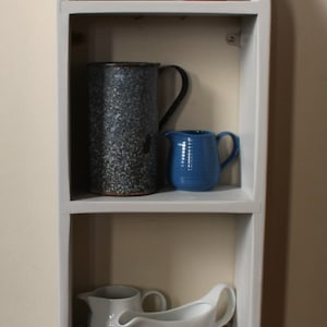 May include: A white painted wooden shelf unit with three shelves. The top shelf has a black heart-shaped chalkboard with the text "Other colours available". The middle shelf has a blue and white speckled pitcher and a blue ceramic pitcher. The bottom shelf has a white ceramic pitcher and a white ceramic gravy boat.