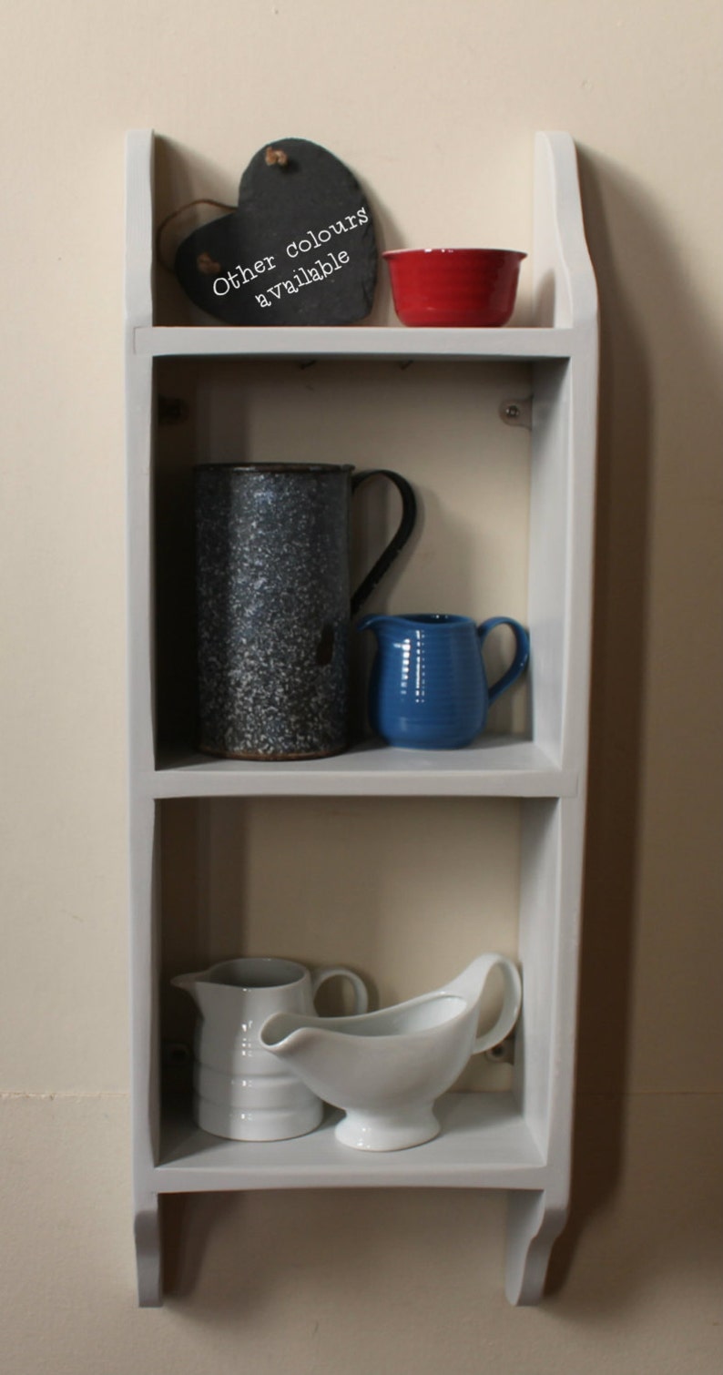 May include: A white painted wooden shelf unit with three shelves. The top shelf has a black heart-shaped chalkboard with the text "Other colours available". The middle shelf has a blue and white speckled pitcher and a blue ceramic pitcher. The bottom shelf has a white ceramic pitcher and a white ceramic gravy boat.