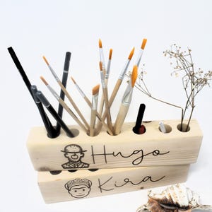 May include: Wooden pencil holder with eight slots, featuring two personalized names, "Hugo" and "Kira", with a cartoon image of a person next to each name. The holder is filled with paintbrushes and pencils.