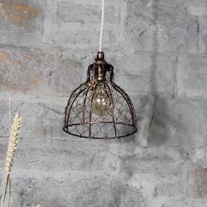 Könnte beinhalten: Eine braune Drahtkäfig-Pendelleuchte mit einer klaren Glühbirne, die an einem weißen Kabel an einer grauen Backsteinwand hängt.