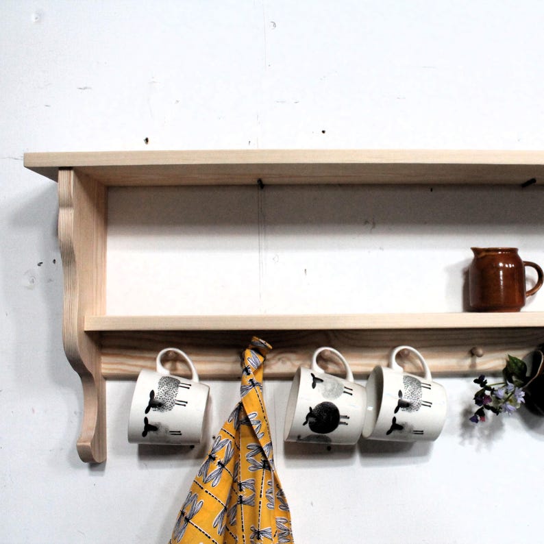 Country style wooden wall shelf with small pegs for mugs on a white wall.  Shown with 3 blck and white mugs.