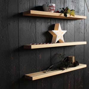 May include: Three light-coloured wooden floating shelves mounted horizontally. The top shelf holds a white bowl with the text Holly Jolly Christmas and holly sprigs. The middle shelf features a white star ornament. The bottom shelf holds a pine branch.