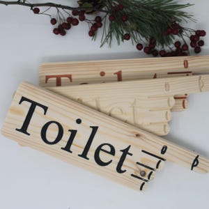 May include: Wooden toilet sign with black lettering. The sign is light-colored with a directional arrow. Other wooden signs with the numbers 0 and 1 are visible in the background. A sprig of evergreen and red berries are in the background.