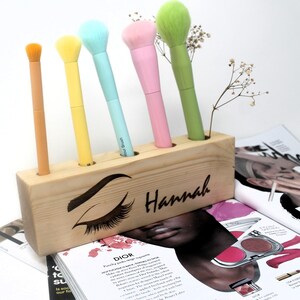 May include: Wooden makeup brush holder with five slots, personalized with the name "Hannah" and a stylized eye design. The holder is filled with five colorful makeup brushes.
