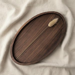 May include: An oval-shaped dark brown wooden tray with a smooth, recessed surface and a raised rim. A single, slender, light beige dried grass stem with a fluffy, plume-like seed head is positioned diagonally across the centre of the tray.