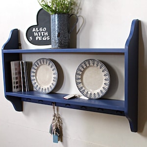 May include: A blue wooden shelf with two shelves and a bottom row of hooks. The top shelf has two decorative plates with a blue and white pattern. The bottom shelf has a stack of books and a small chalkboard sign that says "ALSO WITH 3 PEGS".