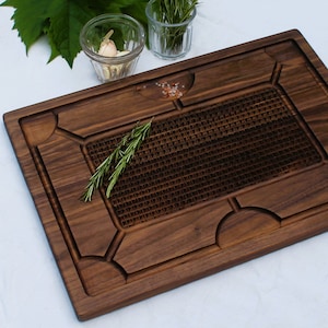 May include: A large, rectangular, dark brown wooden chopping board with a raised, textured grid pattern in the centre. The board has decorative carvings around the edges and a small amount of pink Himalayan salt on the surface.