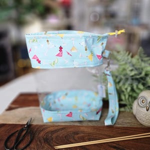 May include: A light blue fabric knitting project bag and a matching rectangular container, both adorned with a cocktail-themed pattern. The bag has a clear plastic window and a wrist strap. A pair of scissors and knitting needles are also visible.