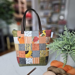 Puede incluir: Un bolso tipo tote de patchwork con una variedad de cuadrados de tela con motivos otoñales en tonos naranja, amarillo, marrón y gris, con asas de cuero marrón oscuro, cierre de cremallera y una pequeña etiqueta blanca en la costura lateral.