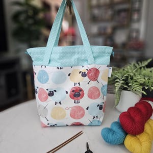 May include: A fabric tote bag featuring a white background with a repeating pattern of colorful sheep and yarn balls. The bag has light blue handles and a matching light blue top section with a white polka dot print. It includes a drawstring closure.