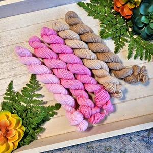 May include: A wooden tray displays several skeins of yarn in shades of pink and tan. The yarn is arranged in neat rows, with the pink hues ranging from light to vibrant. Green fern and faux succulents add a touch of natural color.